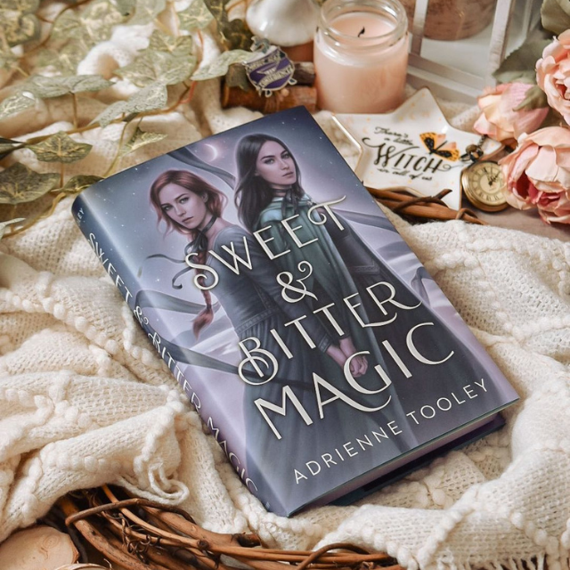 A copy of Sweet & Bitter Magic laying on a white cloth surrounded by vines, flowers, a star shaped trinket dish and a pink candle.