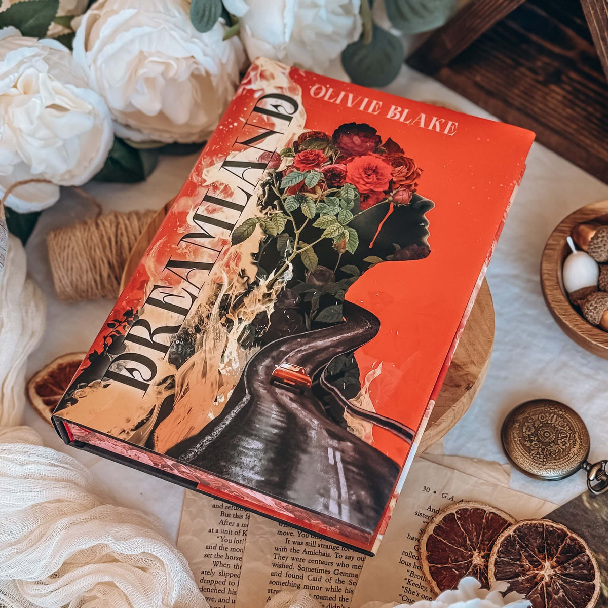 The front cover for the book Dreamland by Olivie Blake featuring an illustrative depiction of roses, flames, and a roadway merging into a face silhouette on a red background. The book is styled on a wooden pedestal surrounded by decorative items.
