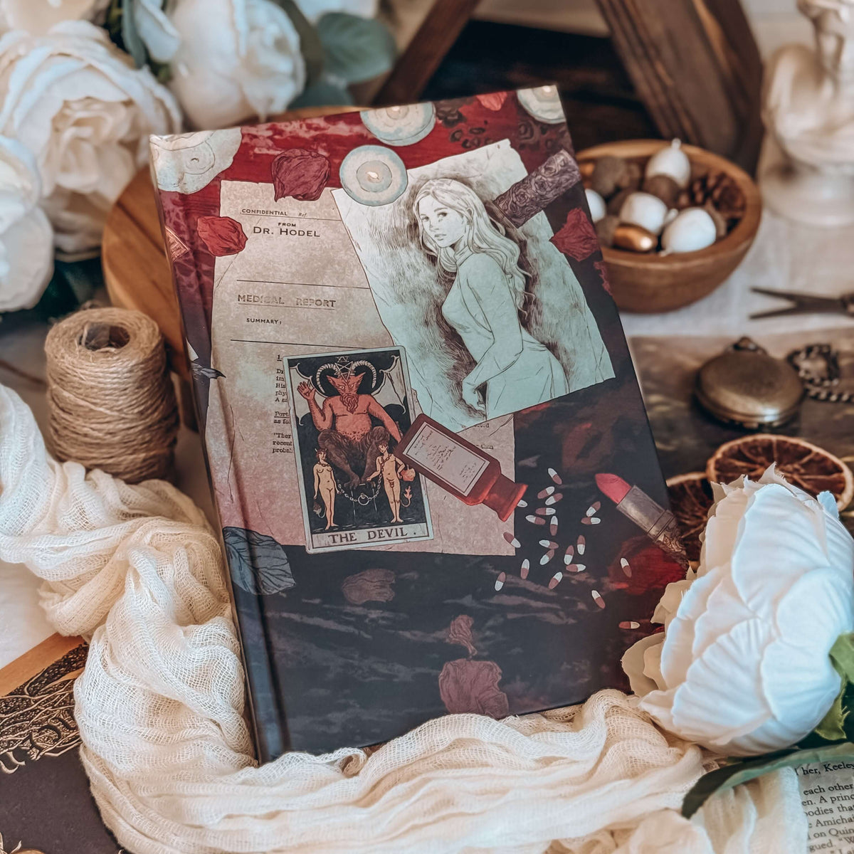 The front hardcover for the book Dreamland by Olivie Blake showing an illustrated collage with a sketch of a woman, a medical report, a tarot card, a spilled bottle of pills. The book rests on wooden pedestal surrounded by white fabric, twine, white flowers, and other decorative items.