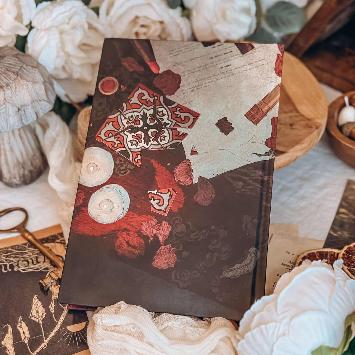 The back hardcover for the book Dreamland by Olivie Blake showing an illustrated collage featuring a medical report, a broken mandela tile, and tealight candles. The book rests on wooden pedestal surrounded by white fabric, twine, white flowers, and other decorative items.