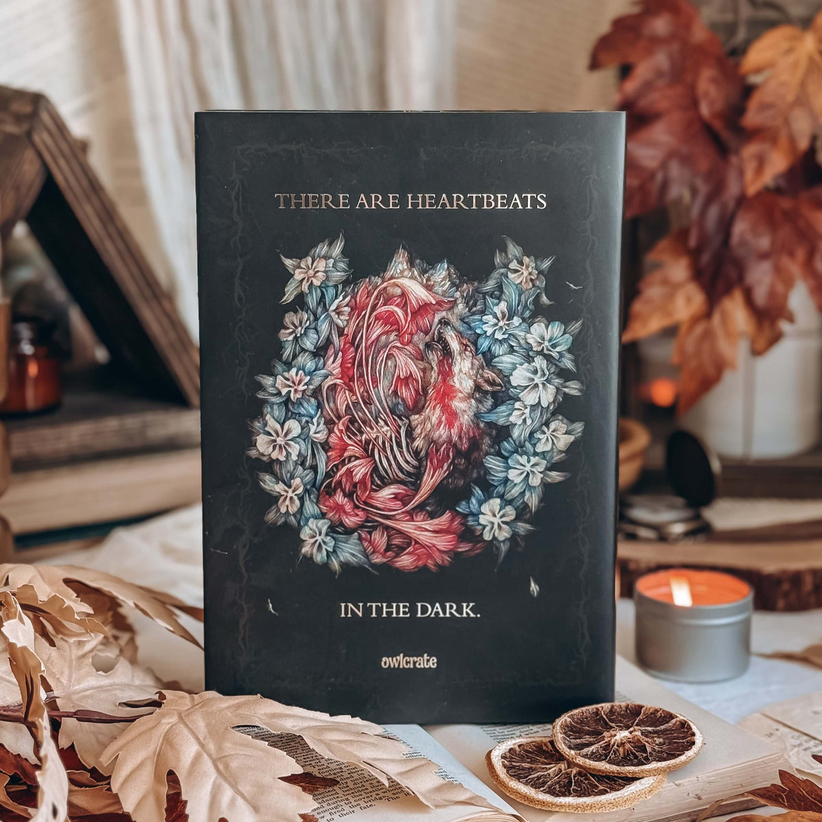 The back cover of a hardcover book featuring a central illustration of a dead animal with glowing flowers emerging from its exposed ribcage, surrounded by blue and white flowers. The quote 'There are heartbeats in the dark' surrounds the image in gold foil font.