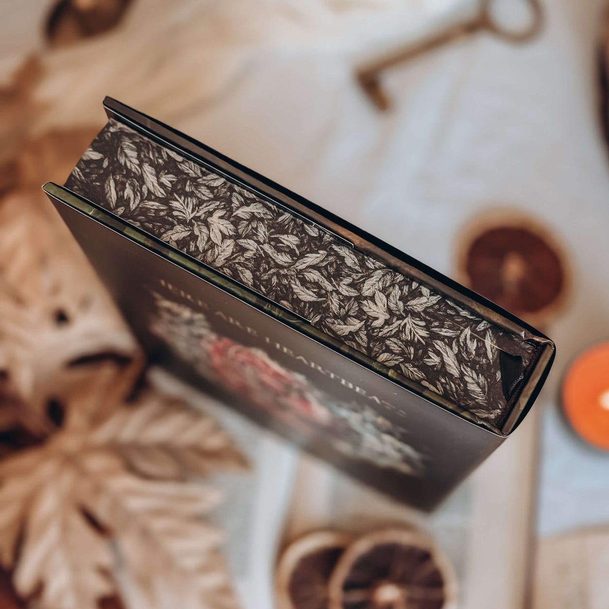 A close-up of the illustrated stenciled edges of a hardcover book is featured showing detailed black-and-white artwork of leaves. The out-of-focus background shows various autumn-themed decorative items.