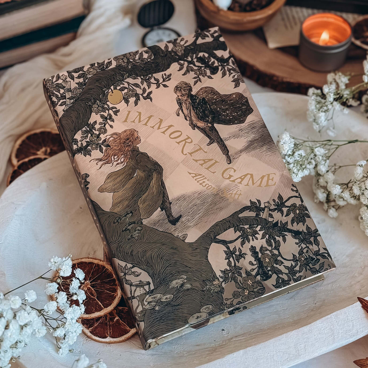 A hardcover book titled Immortal Game by Allison Saft with an illustrated cover featuring a man and woman framed by a flowering tree branch. Book is surrounded by various decorative items.