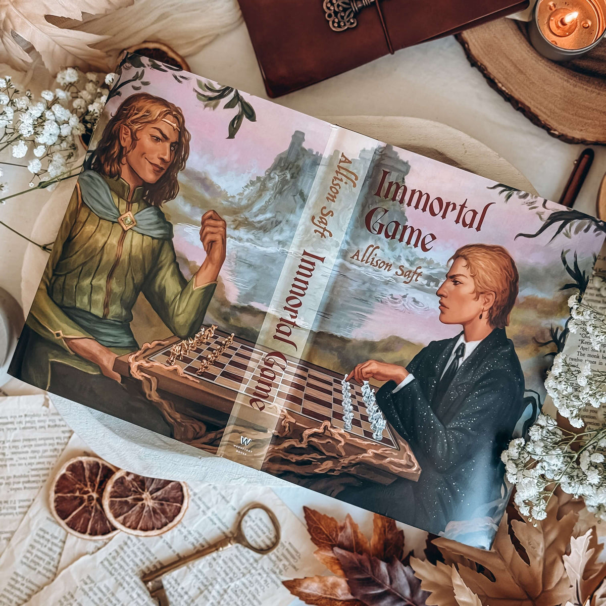 A flat-lay showing the reversible dust jacket for Immortal Game by Allison Saft surrounded by autumnal-themed decorative items. The dust jacket artwork shows an illustration featuring two characters, one in contemporary clothing and the other in fantasy clothing, playing chess.
