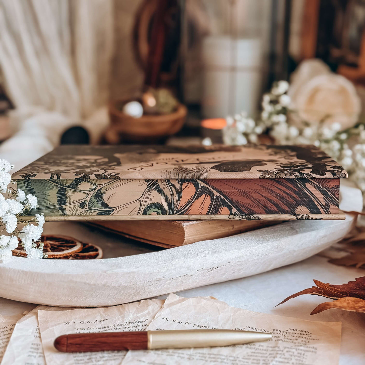 The detailed stencilled edges of a hardcover book sits on a white tray surrounded by various vintage-inspired decor. The stencilled edges resemble a vintage close-up illustration of a butterfliy's wing in shades of orange, tan and green.