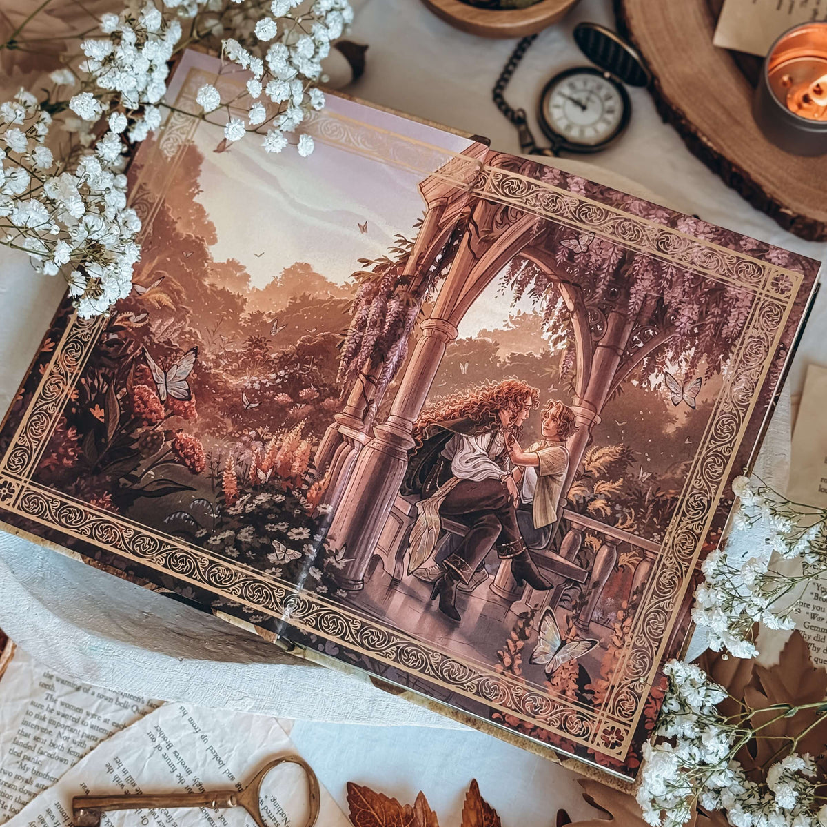 An open book on a white cloth background featuring baby's breath flowers and various decorative items. The endpapers feature a detailed fantasy illustrated scene of a couple under an arched pavilion surrounded by flowers and butterflies.