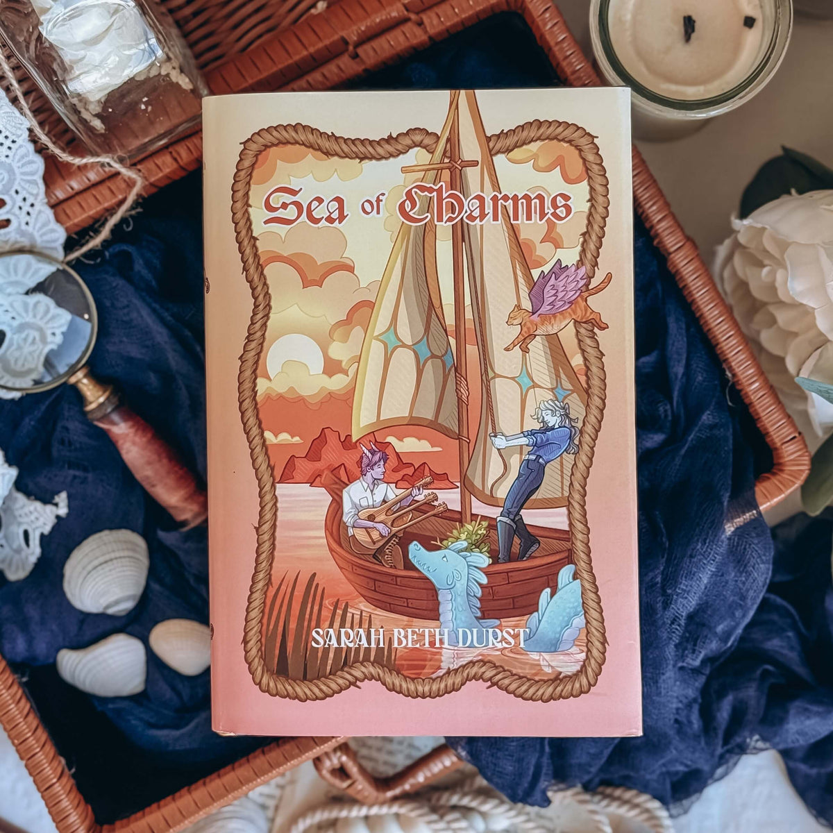 The front reversible dust jacket for the book Sea of Charms by Sarah Beth Durst shown resting on an open wicker basket with a dark blue fabric and sea shells. The illustration shows two characters on a small sailboat with a blue sea creature floating by.