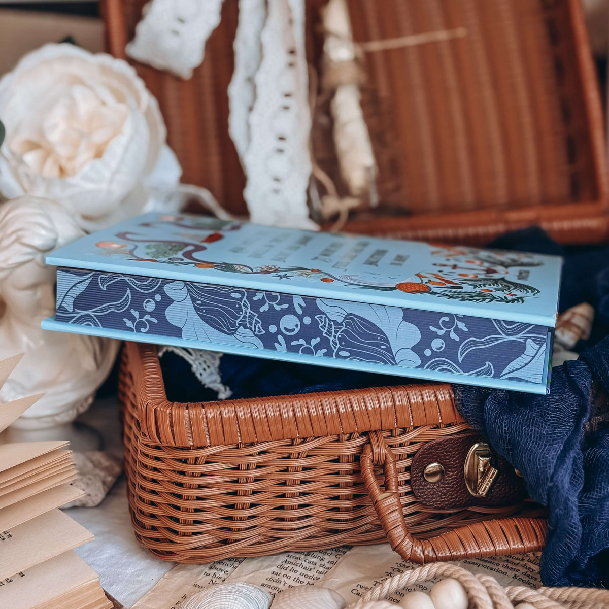 The stenciled edges for the book Sea of Charms by Sarah Beth Durst styled in an open wicker basket surrounded by decorative items. The nautical-inspired illustrated edges depict kelp and seashells on a dark blue background.