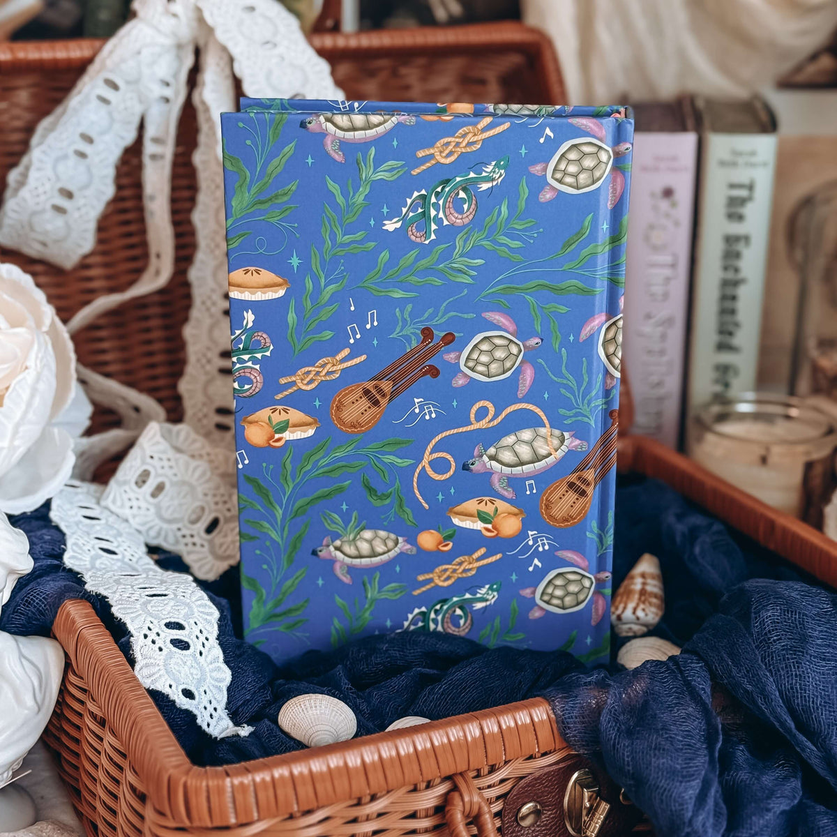 The hardcover for Sea of Charms by Sarah Beth Durst is feautres a whimsical ocean-themed illustration of sea turtles, kelp, and musical instruments, displayed inside a wicker basket with lace and fabric.