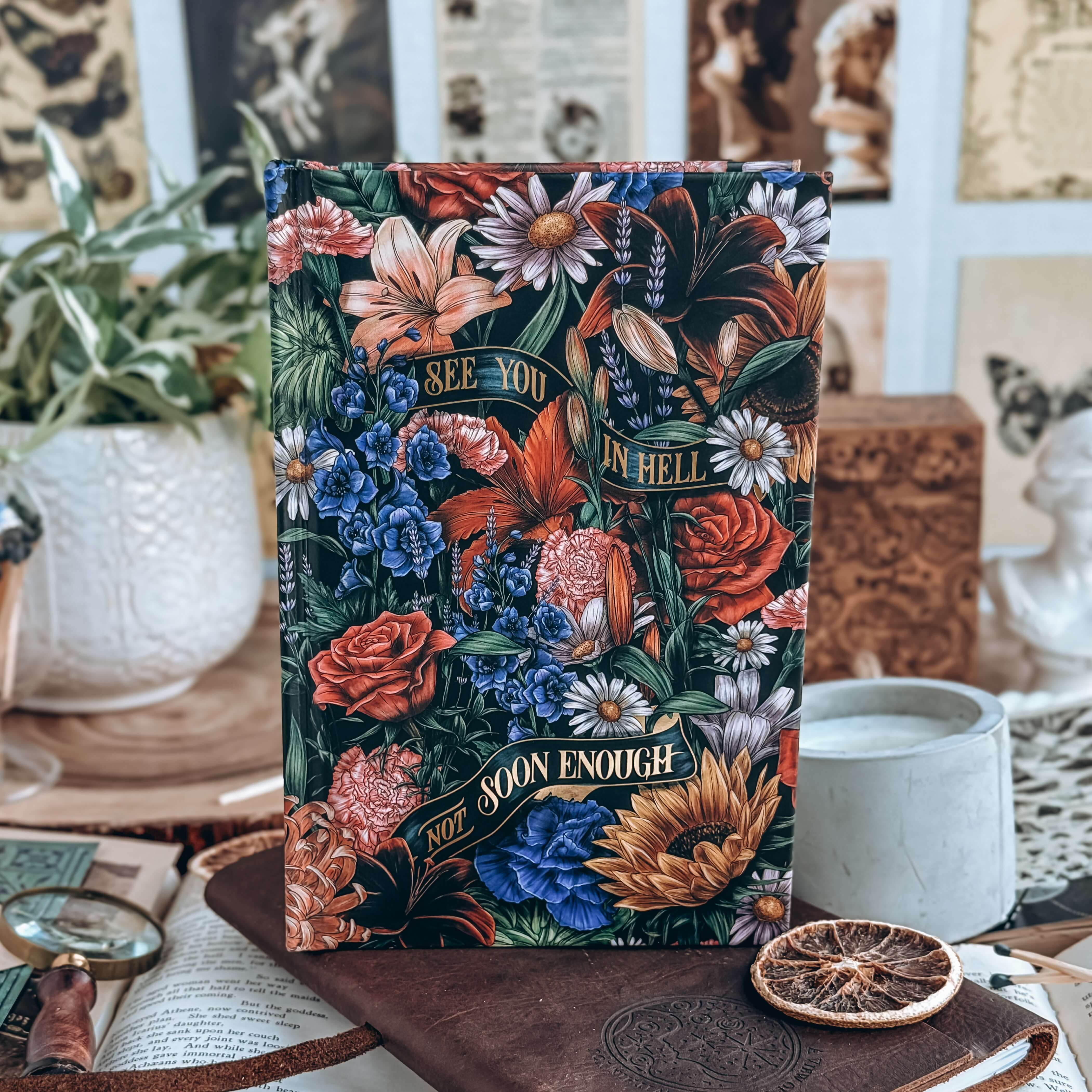 Hardcover of Starter Villain by John Scalzi. Decorated floral book includes text reading "See you in Hell. Not Soon enough." Book is surrounded by decorative items.