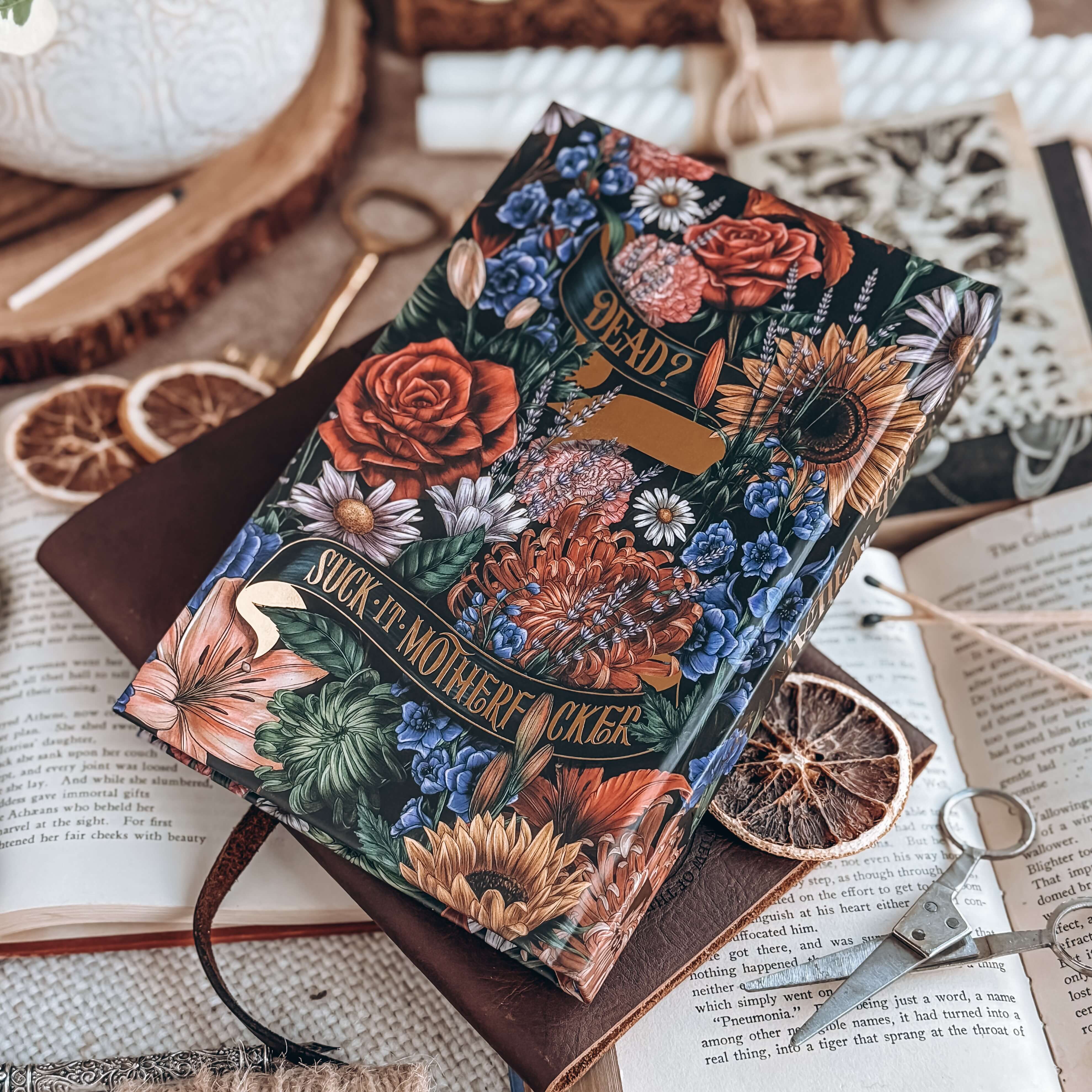 Hardcover of Starter Villain by John Scalzi. Decorated floral book features text reading "Dead? Suit it, motherfucker." Book is surrounded by decorative items.