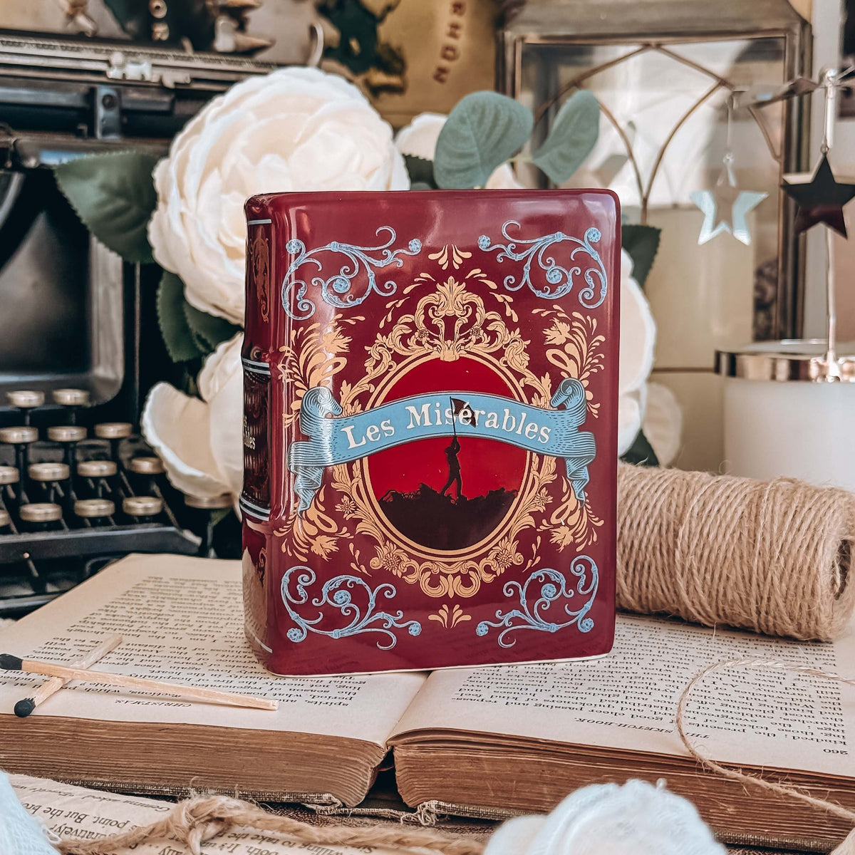 The front of a book-shaped vase modeled after a copy of Les Miserables sits on an open book with twine, white flowers, and a typewriter in the background.