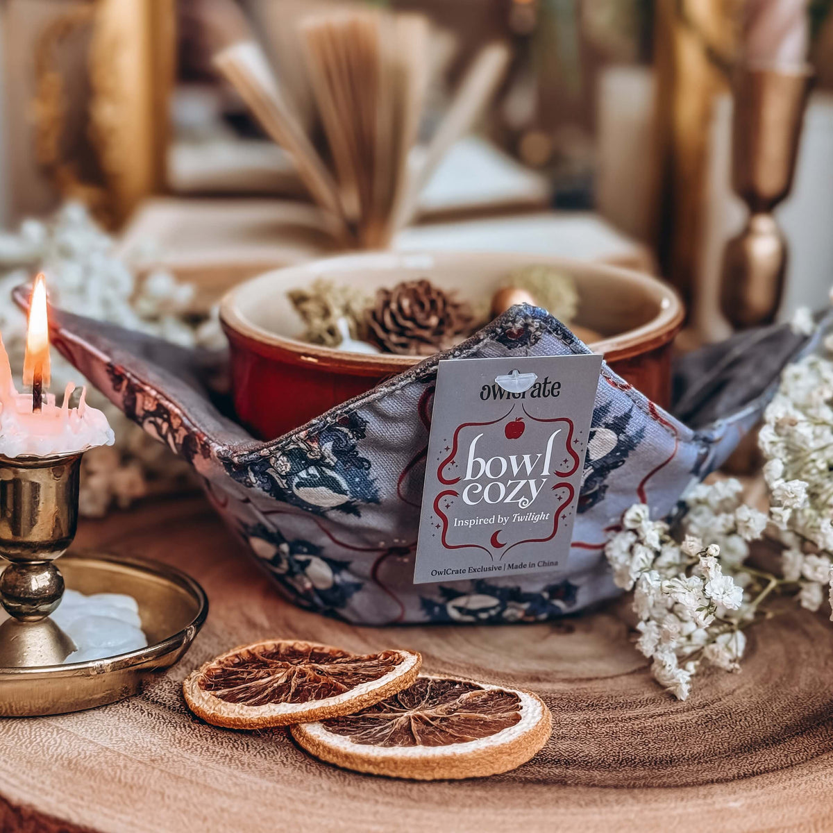 A grey fabric bowl cozy is featured with a grey tag prominently shown. A red bowl of potpouri rests inside the bowl cozy with dried orange slices, a lit candle, baby's breath flowers, and an open book surrounding.