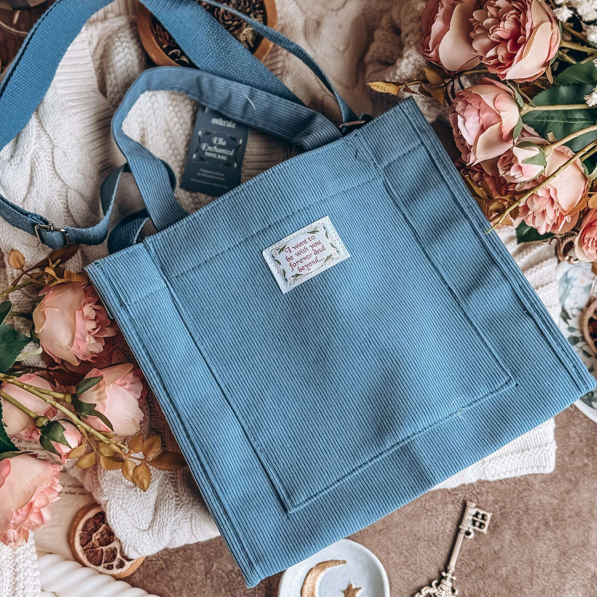 Ella Enchanted-inspired blue corduroy tote bag lies on white knit sweater surrounded by pink flowers.