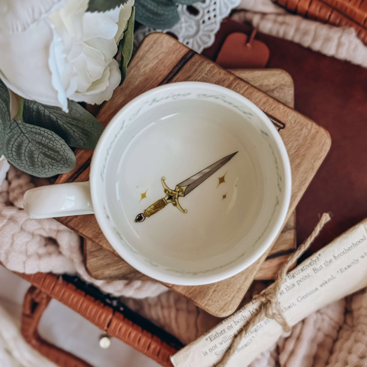 The topview of teacup with a dagger printed on the buttom of the cup sits on a stack of wooden coasters. The cup is surrounded by cream fabrics and white flowers.