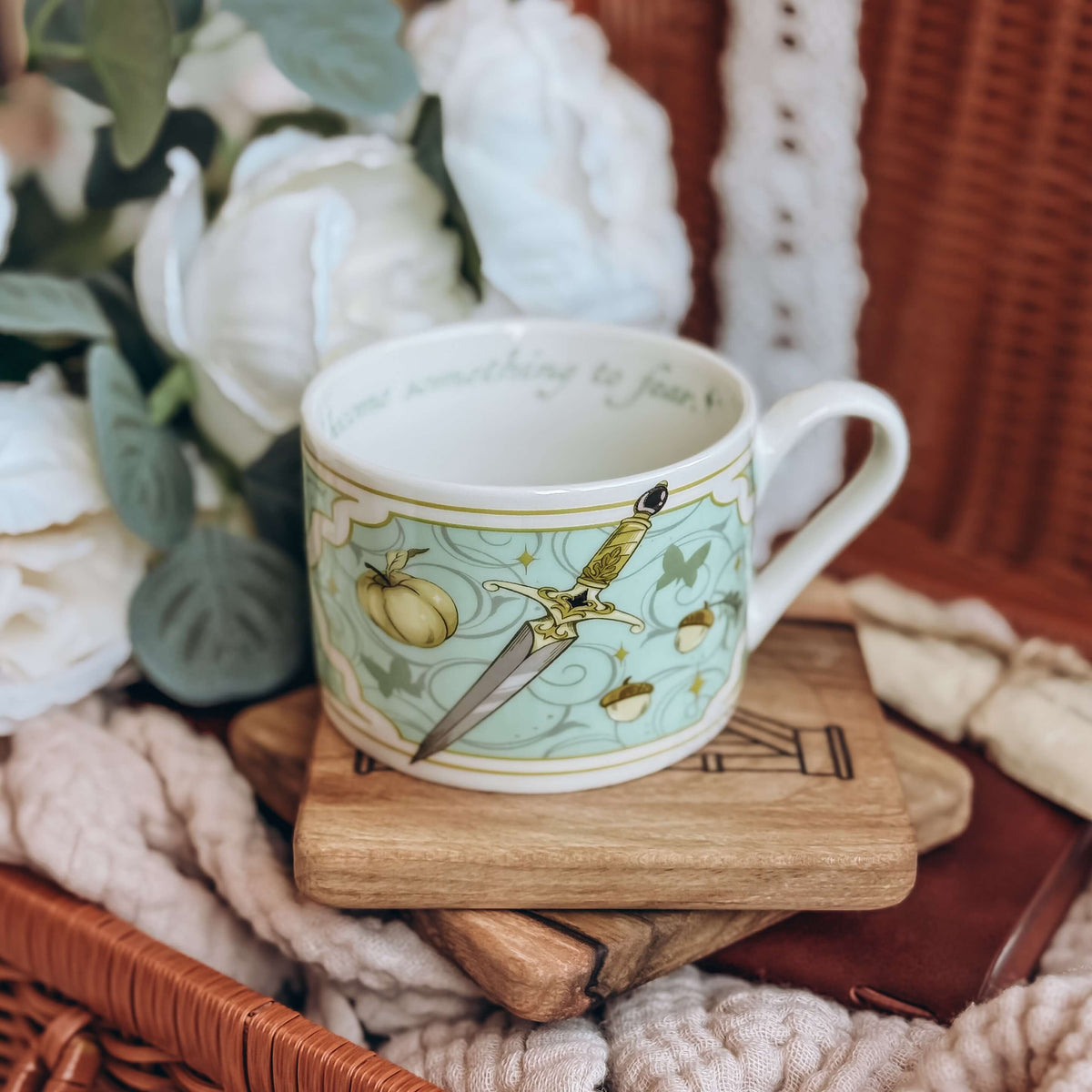 A teacup featuring a motif of daggers and acorns on a light teal background sits on a stack of wooden coasters. The cup is surrounded by cream fabrics and white flowers.