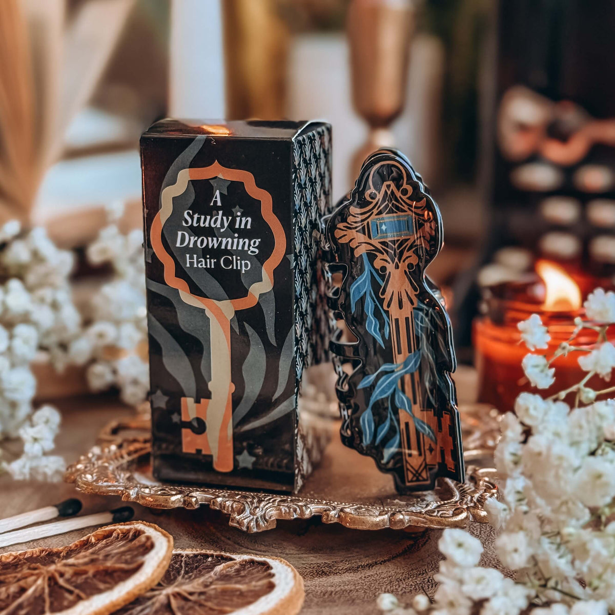 A hair clip that resembles a skeleton key wrapped in kelp leans against a box with the text A Study in Drowning hair clip printed on it. The hair clip and the box sit on a orange dish surrounded by dried orange slices, baby's breath flowers, and a lit candle.
