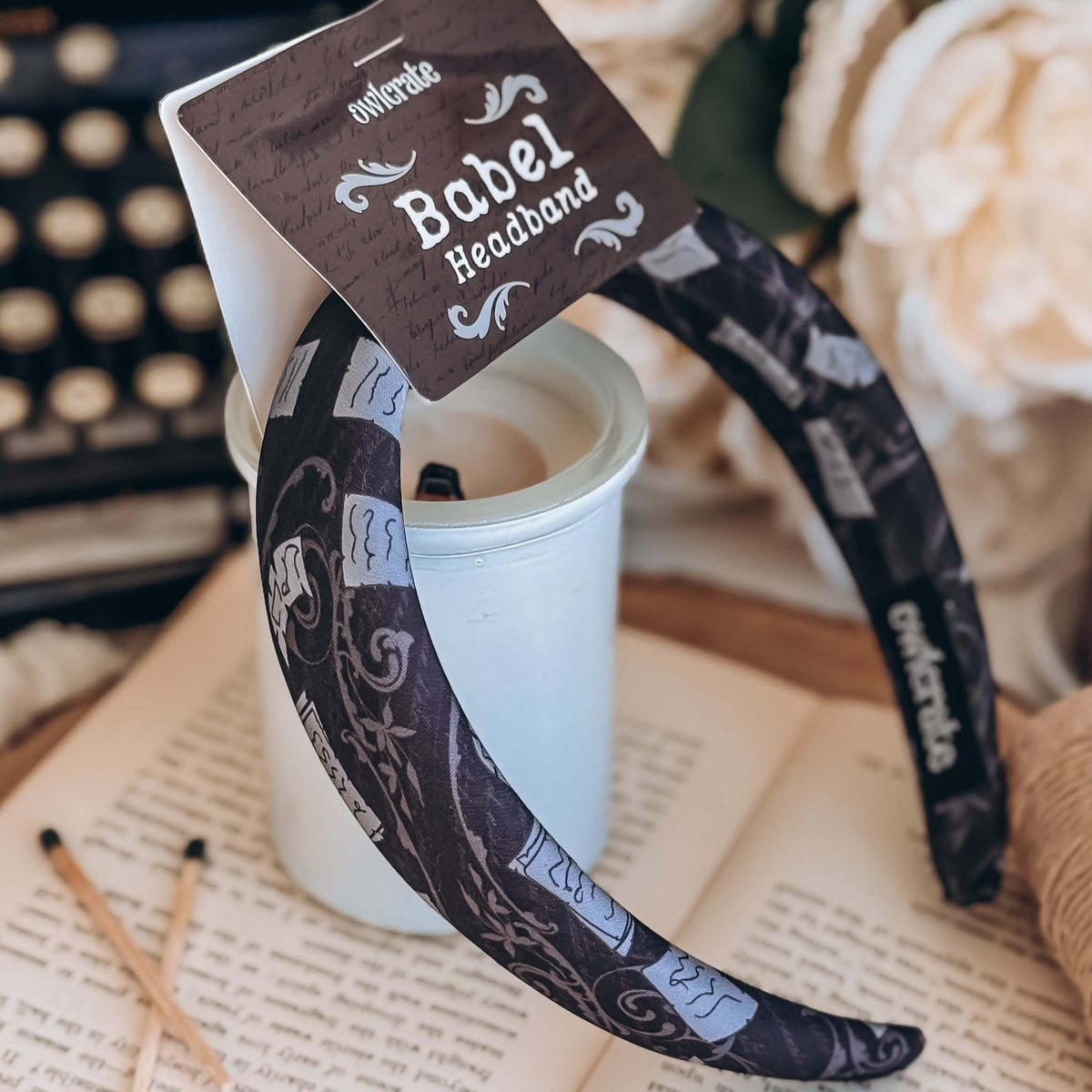 A black fabric headband with a pattern made of pages and decorative designs rests on an unlit candle votive.
