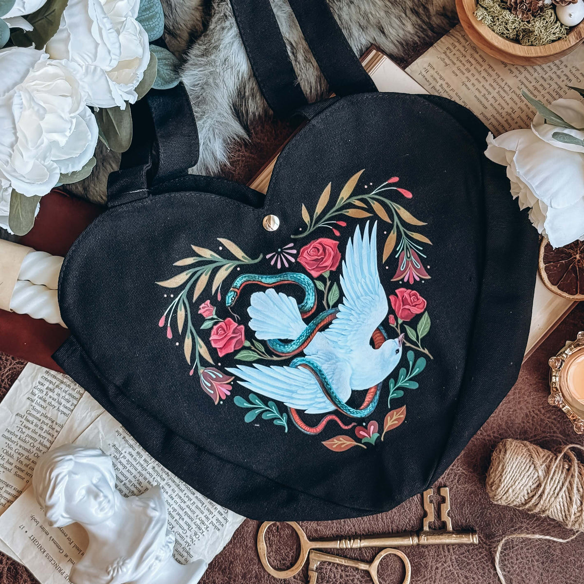 A black heart-shaped tote back featuring a a dove and serpent design is centered with keys, marble busts, books, twine, and white flowers surrounding.
