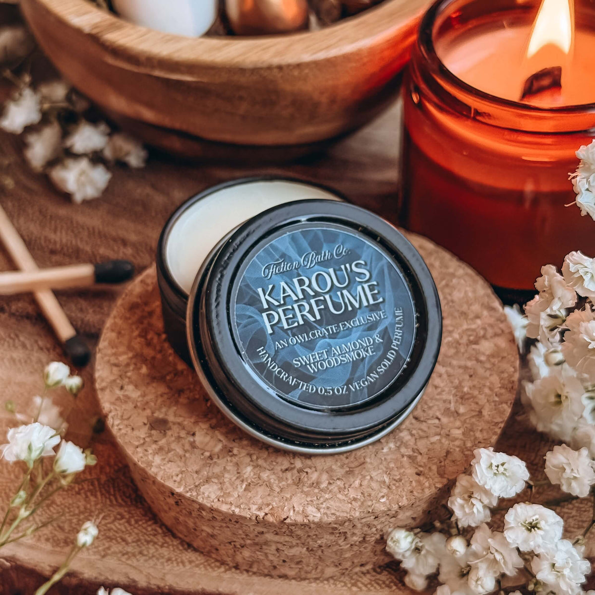 A small open tin of Karou's Perfume sits on a cork stand, with baby's breath flowers, a lit candle, and two matchsticks in the background.
