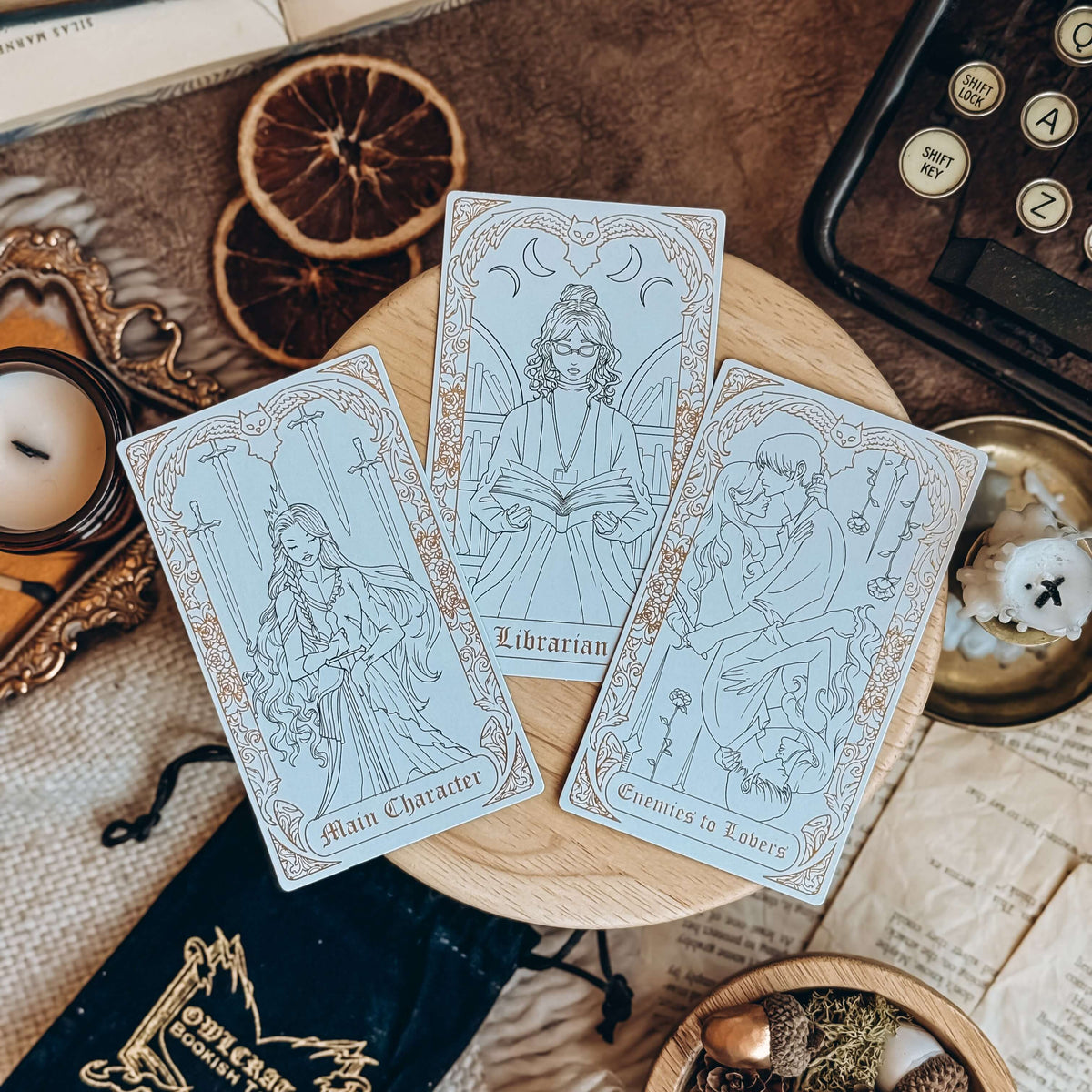 Three gold-foiled tarot cards, depicting the Main Character, Librarian, and Enemies to Lovers, lay on a wooden stand surrounded by candles, a typewriter, and papers.
