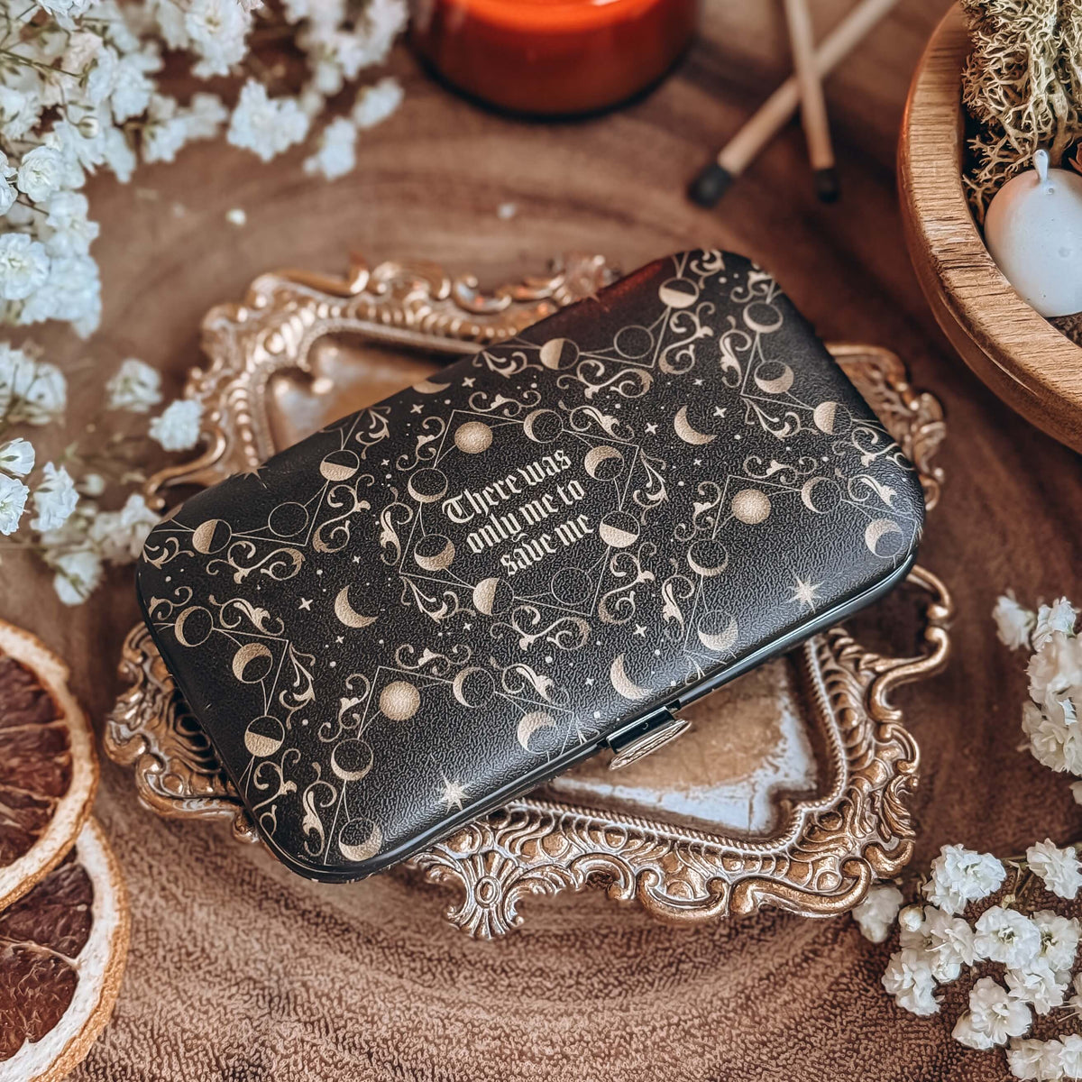 A black case with celestial designs and the quote There was only me to save me is centered, with baby's breath flowers, potpouri, and matchsticks surrounding.