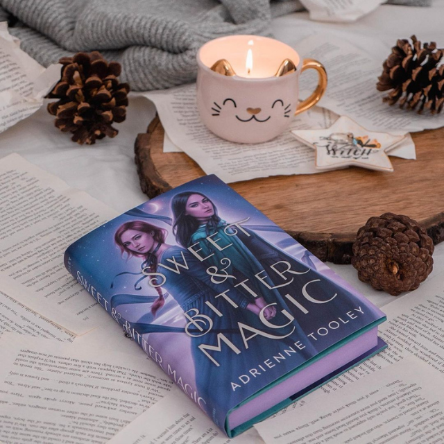 A copy of Sweet & Bitter magic laying on some loose book pages beside a wooden try with a cat shaped candle and a star shaped dish on it.