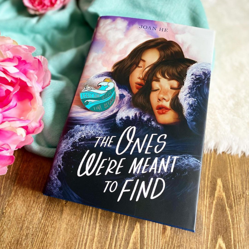 A copy of The Ones We're Meant to Find laying half on a while rug, half on a wooden floor with a piece of green fabric and some pink flowers beside it.