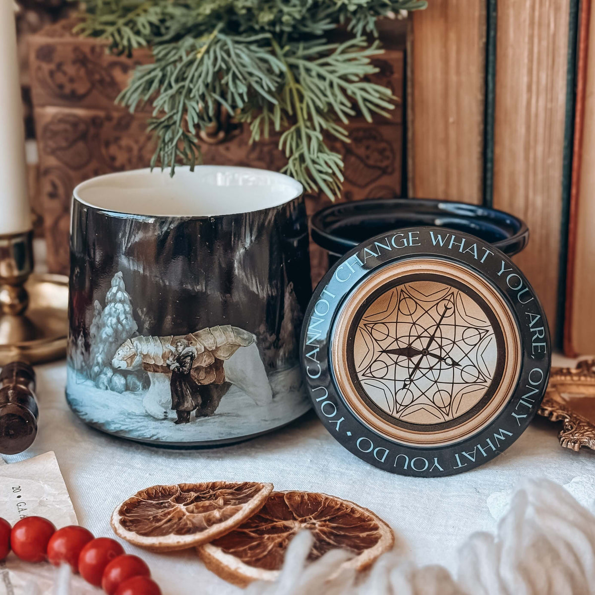 A ceramic mug sits next to a matching ceramic lid, surrounded by various holiday-themed decorative items. The mug features artwork showing an armoured bear next to a small girl in a snowy landscape. The lid features a the quote 'You cannot change what you are, only what you do' circling a fantasy gold-coloured compass.