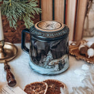 A ceramic mug with a lid surrounded by various holiday-themed decorative items. The mug features artwork showing an armoured bear next to a small girl in a snowy landscape.