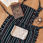 The Night Circus-inspired apron lies on a wooden floor next to a book and a candle.