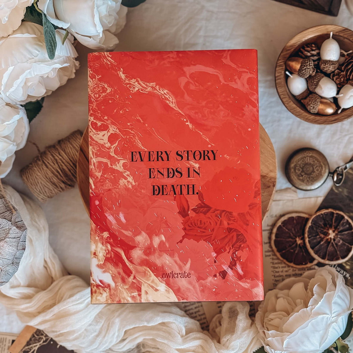 The back cover for the book Dreamland by Olivie Blake showing a red and orange marbling effect that merges into flames and illustated roses with the quote 'Every story ends in death' centered. The book is styled on a wooden pedestal surrounded by decorative items.