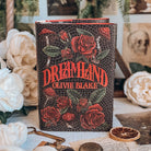 The front reversible dust jacket for the book for the book Dreamland by Olivie Blake showing a dark mosaic pattern featuring roses and mushrooms with the title and author name in large red font. The book stands upright surrounded by a variety of decorative items, such as white flowers, and white papers. 