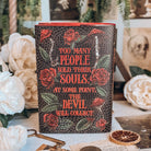 The back reversible dust jacket for the book for the book Dreamland by Olivie Blake showing a dark mosaic pattern featuring roses and mushrooms with a quote in large red font. The book stands upright surrounded by a variety of decorative items, such as white flowers, and white papers. 