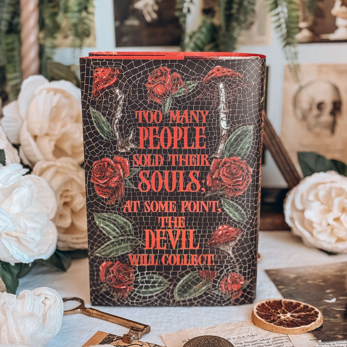 The back reversible dust jacket for the book for the book Dreamland by Olivie Blake showing a dark mosaic pattern featuring roses and mushrooms with a quote in large red font. The book stands upright surrounded by a variety of decorative items, such as white flowers, and white papers. 
