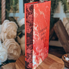 The stenciled edges for the book Dreamland by Olivie Blake are featured, showing a red rose pattern. The upright book is styled on a wooden pedestal surrounded by white flowers, twine and other decorative items. 