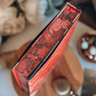 A close-up for the book Dreamland by Olivie Blake showing a red rose pattern. The upright book is styled on a wooden pedestal surrounded by white flowers, twine and other decorative items. 