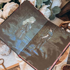 The front illustrated endpapers for the book Dreamland by Olivie Blake showing a man and woman standing in a derelict room. The open book is styled among white fabric, white flowers, and other decorative items. 