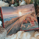 The back illustrated endpapers for the book Dreamland by Olivie Blake showing a man and woman in a red convertible driving along an empty coastal highway at sunset. The open book is styled among white fabric, white flowers, and other decorative items. 