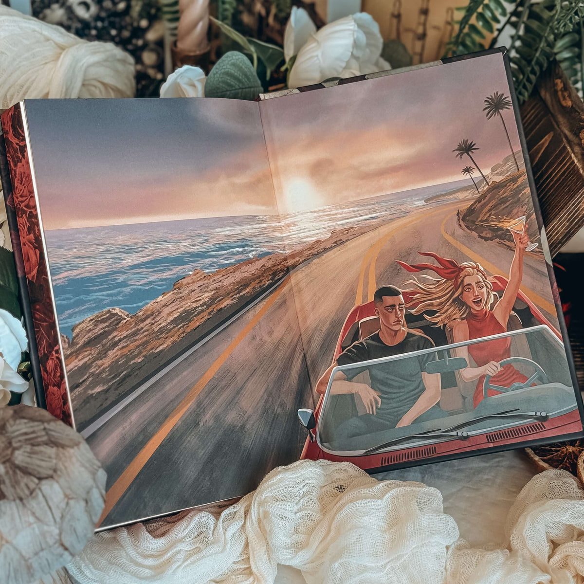 The back illustrated endpapers for the book Dreamland by Olivie Blake showing a man and woman in a red convertible driving along an empty coastal highway at sunset. The open book is styled among white fabric, white flowers, and other decorative items. 