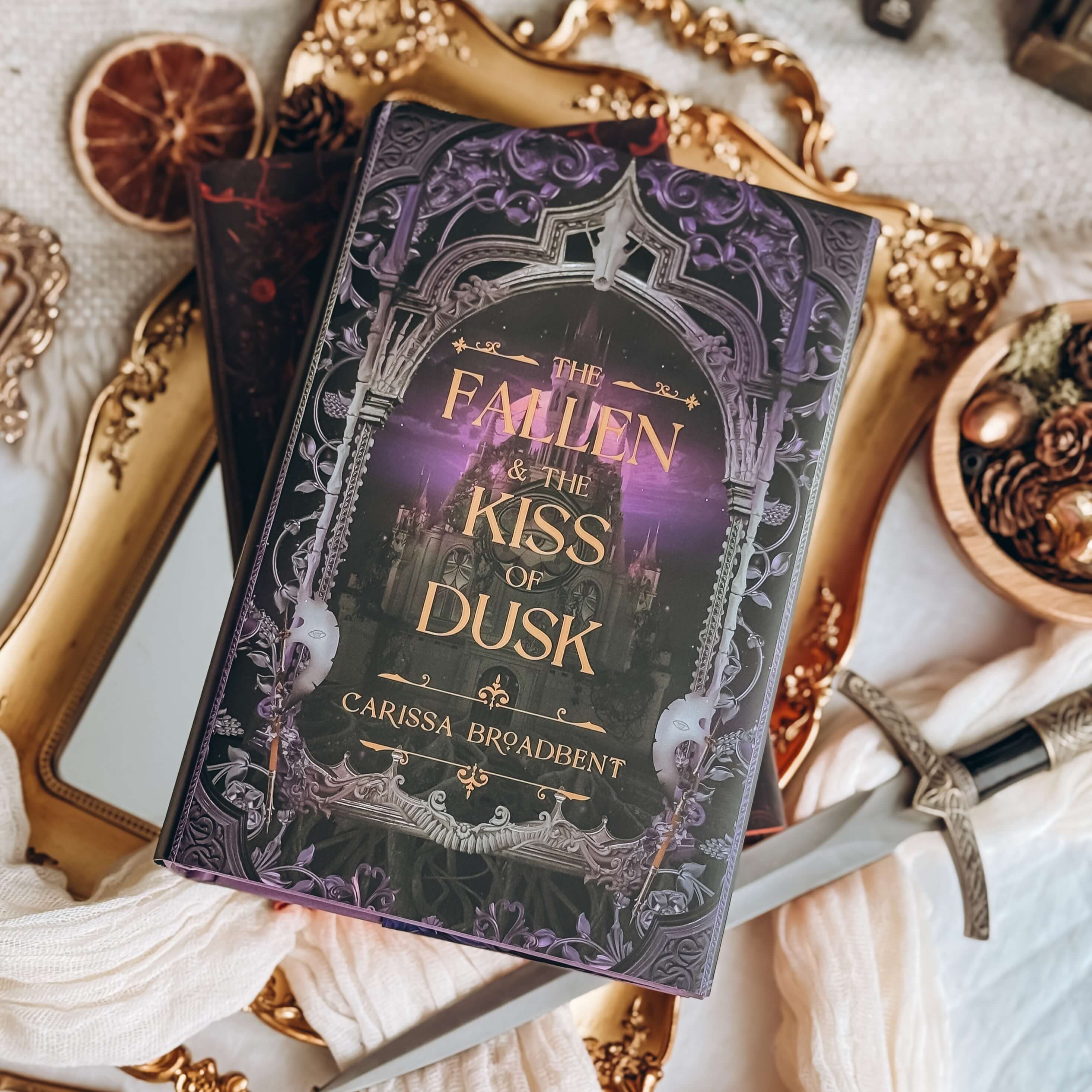 Cover of The Fallen and the Kiss of Dusk by Carissa Broadbent showing a spired building framed by purple-tinted gothic architecture. Book is surrounded by decorative items. 