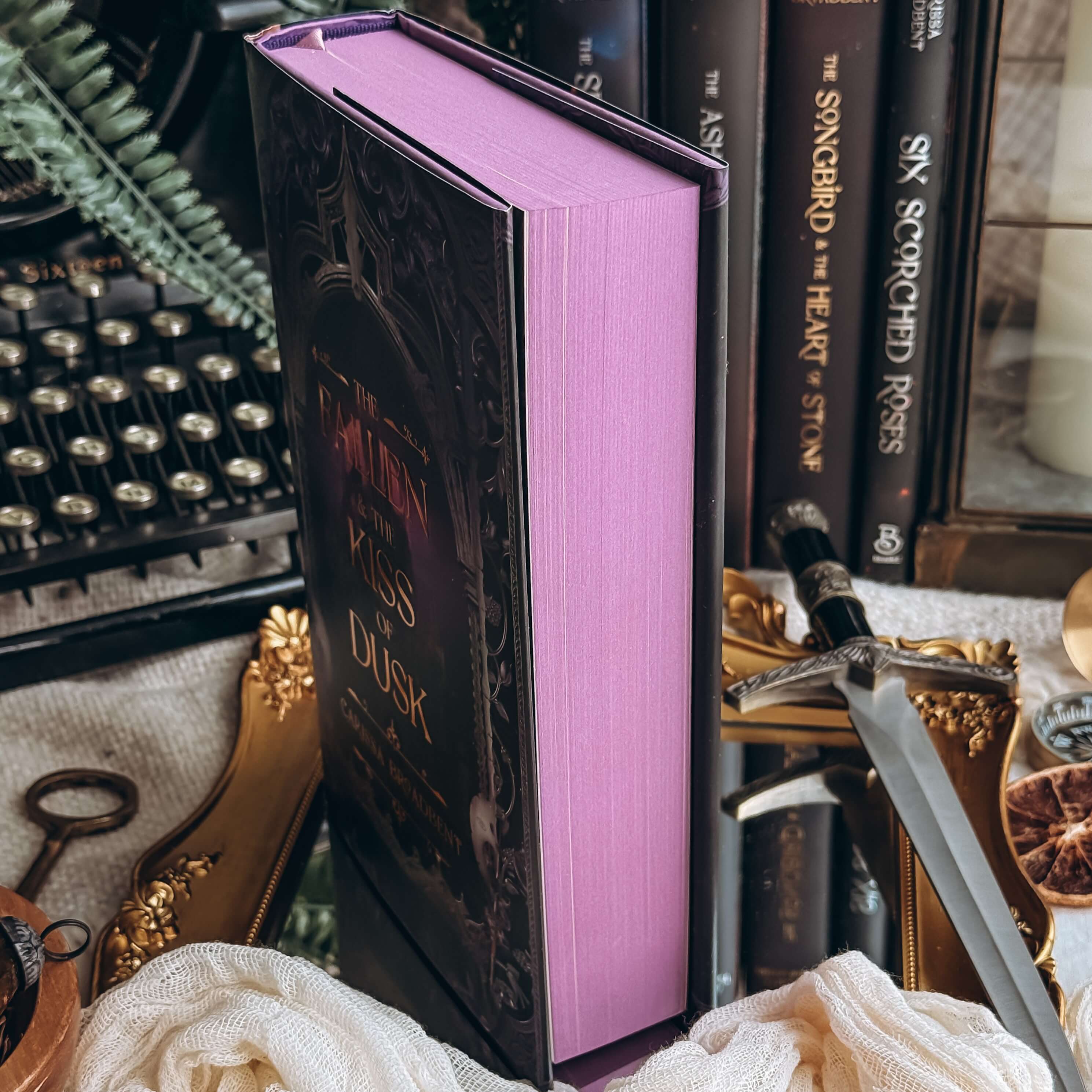 Stained edges of the book The Fallen and the Kiss of Dusk by Carissa Broadbent. Endpapers are a solid purple, the book is surrounded by decorative items and the other books in the series in the background. 