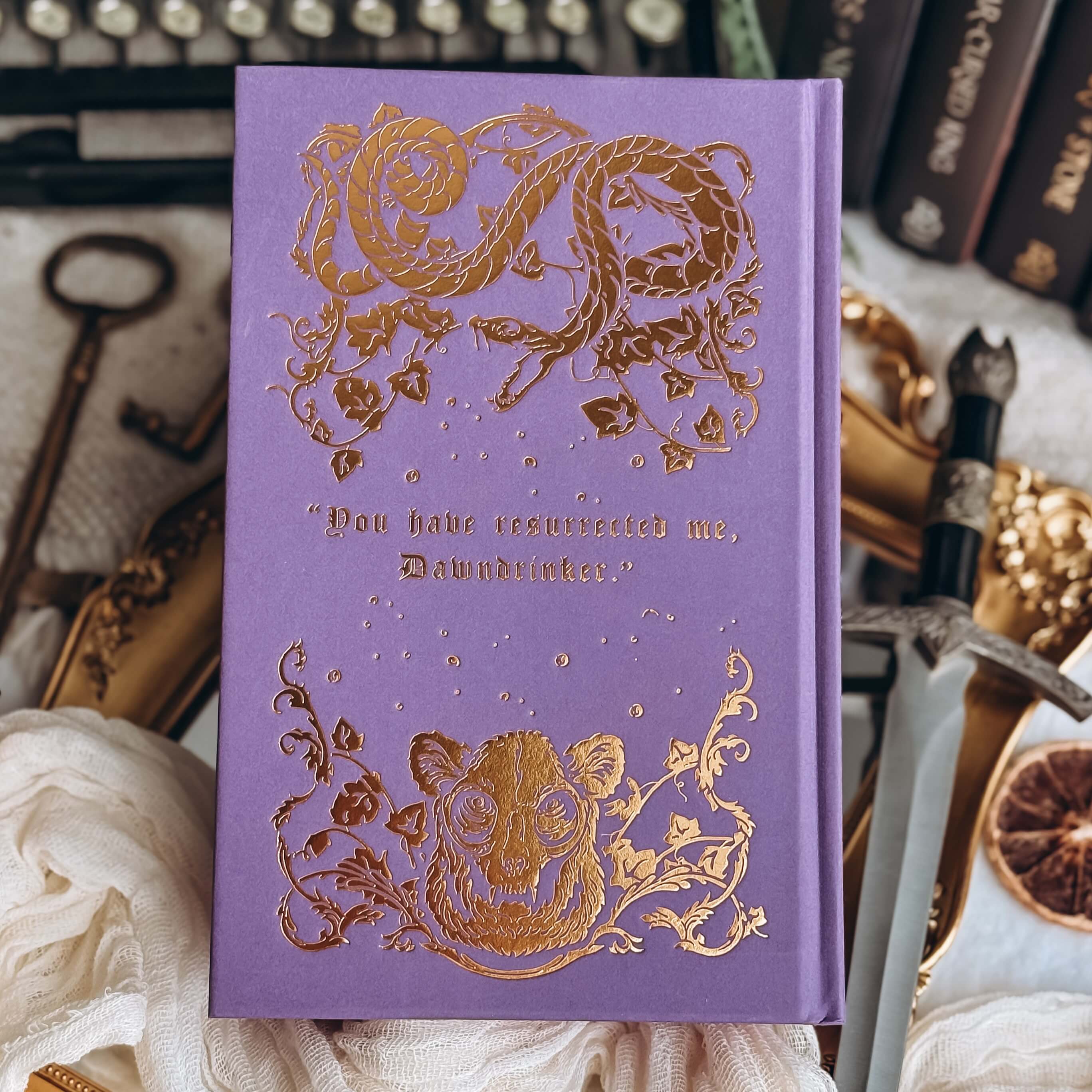 Hardcover of The Fallen and the Kiss of Dusk by Carissa Broadbent. The purple book shows an illustrated snake and furred animal in gold embossing, with the text "You have resurrect me, Dawndrinker."