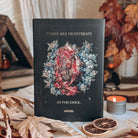 The back cover of a hardcover book featuring a central illustration of a dead animal with glowing flowers emerging from its exposed ribcage, surrounded by blue and white flowers. The quote 'There are heartbeats in the dark' surrounds the image in gold foil font.