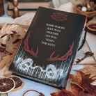 The back cover of a book is centered surrounded by various autumn-themed decorative items. The book design features the quote 'Some places just pull harder on you than the rest' centered on a black background with red antlers, white illustrated trees, and several floating outlines of red eyes.