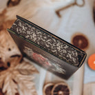 A close-up of the illustrated stenciled edges of a hardcover book is featured showing detailed black-and-white artwork of leaves. The out-of-focus background shows various autumn-themed decorative items. 