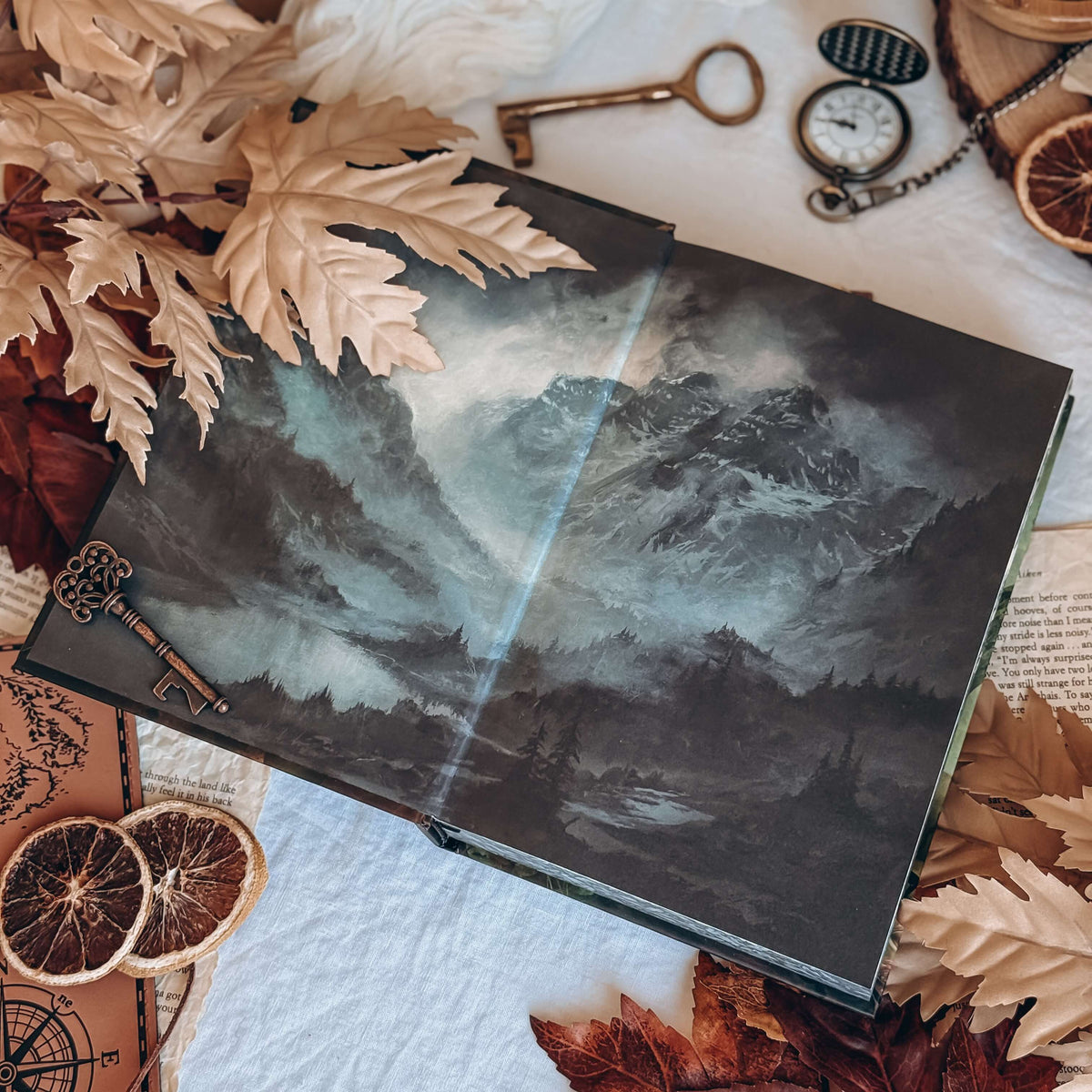 An open book showing the front endpapers depicting a gloomy, misty mountain range. The book is centered surrounded by brown leaves, dried orange slices, antique keys, and a pocket watch.