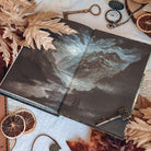 An open book featuring the back endpapers depicting a gloomy, misty mountain range at night. The book is centered surrounded by autumn leaves, dried orange slices, antique keys, and a pocket watch. 