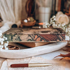 The detailed stencilled edges of a hardcover book sits on a white tray surrounded by various vintage-inspired decor. The stencilled edges resemble a vintage close-up illustration of a butterfliy's wing in shades of orange, tan and green.