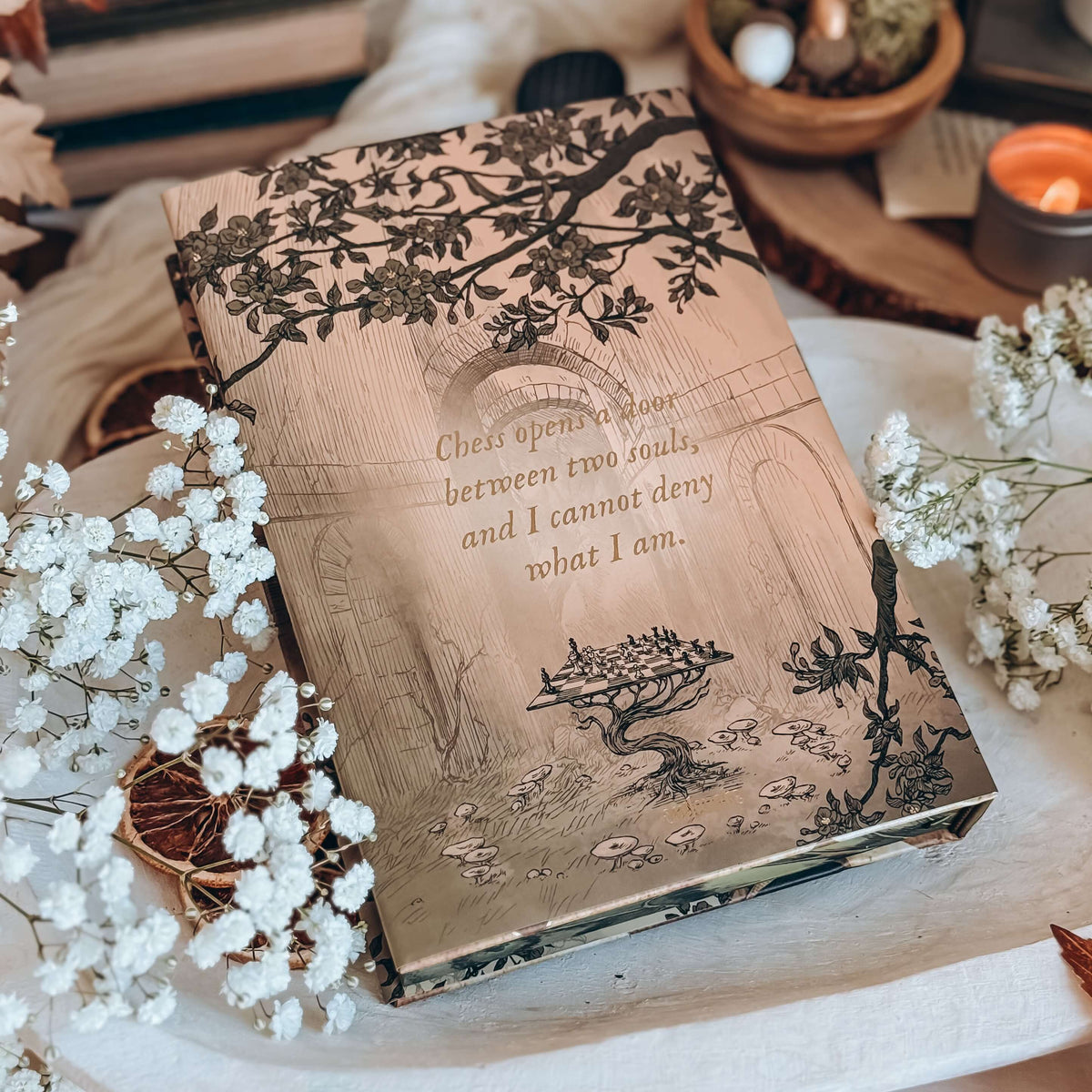 The back of a hardcover book sits on a white tray surrounded by baby's breath flowers, dried orange slices, and other various decor. The illustration of the book features a drawing of a chessboard held up by a sprouting root in an empty stone room, with a quote centered reading 'Chess opens a door between two souls, and I cannot deny what I am.'